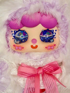 Plush doll face with purple hair and glittery eyes, wrapped in white lace hood