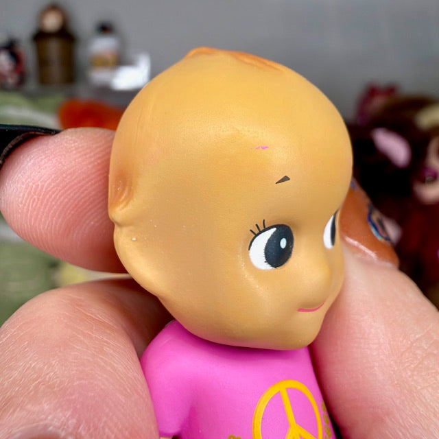 Close-up of pink mini figurine face with large blue eyes and purple peace shirt