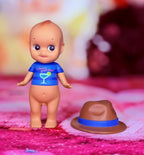 Toy angel figure with blue shirt beside brown fedora hat on pink fabric