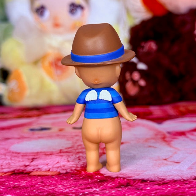 Small toy angel figure with blue shirt and brown hat, standing on pink fabric