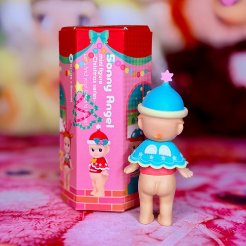 Teal-capped toy figure in blue dress, standing beside Christmas gift box